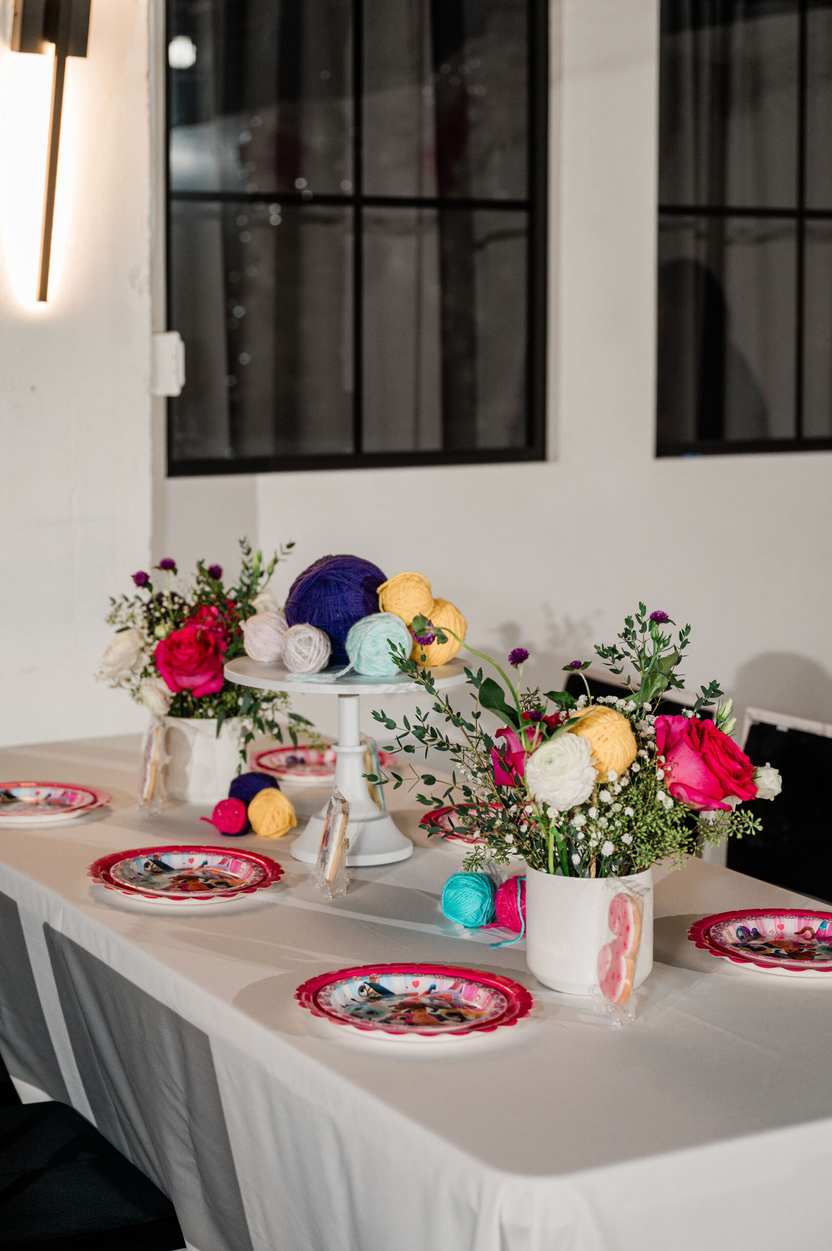 Custom decorated kids party table with themed super kitty cookies and plates, yarn balls and bold pink, white and yellow floral centerpieces with white chairs and white linens.