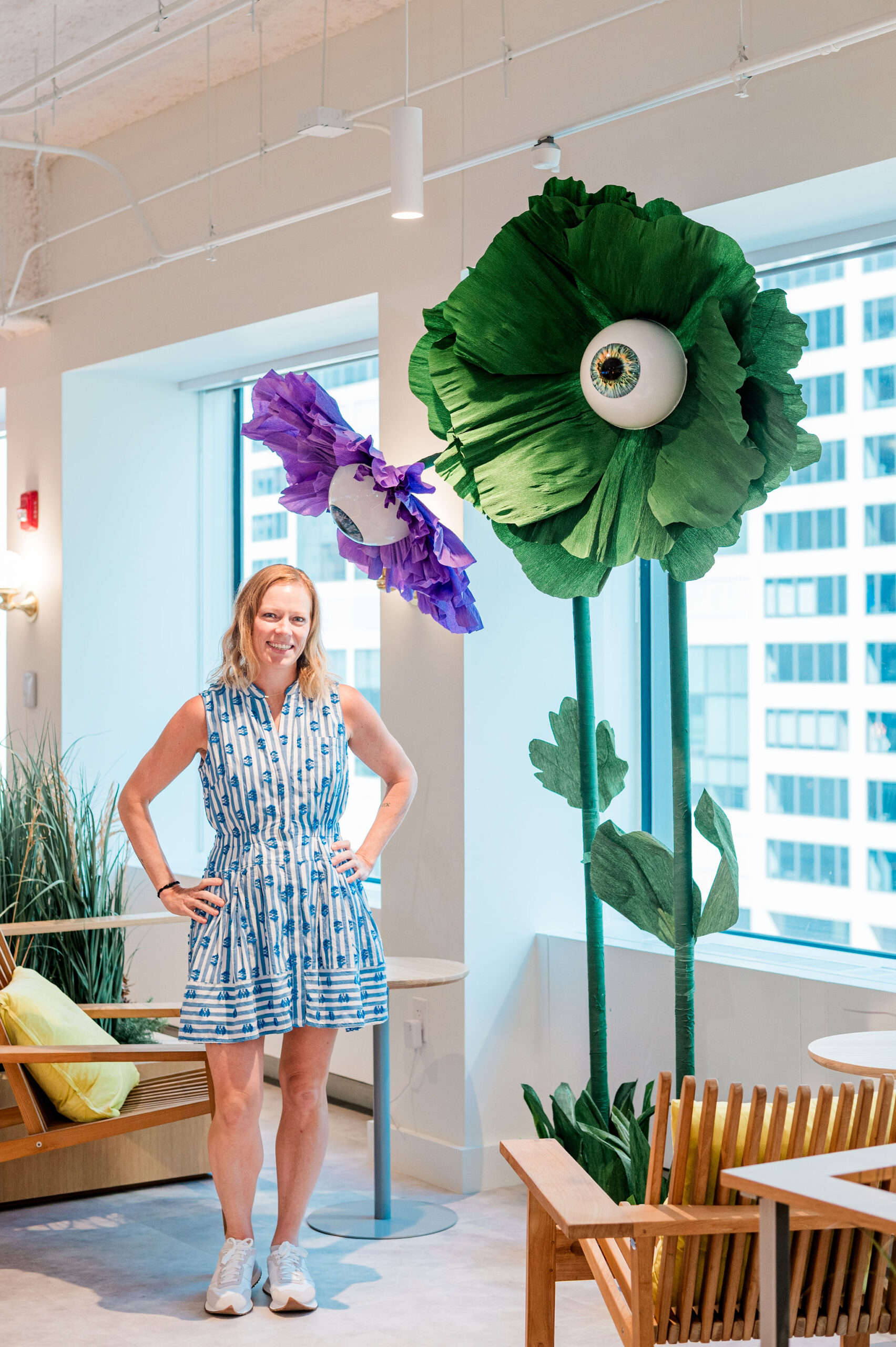 A women in a white and blue dress with her hands on her hips posing in front of two giant surreal eyeball flowers in green and purple
