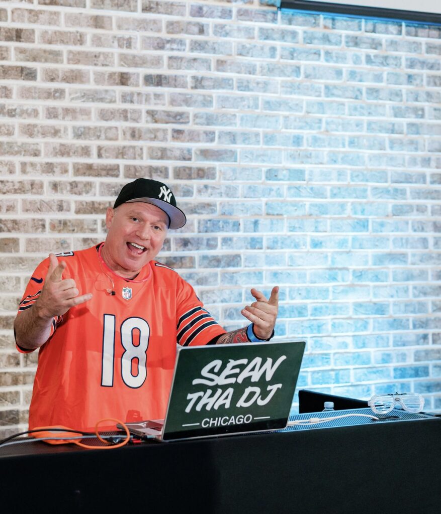 A man in a orange jersey behind a dj booth with a lap top that says sean tha dj.