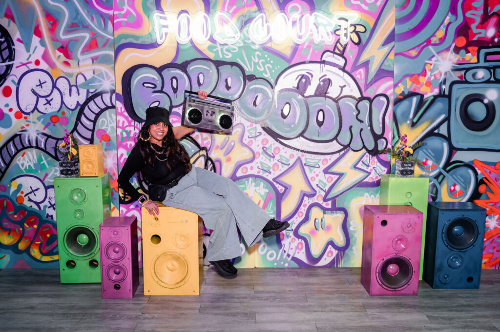 A lady leaning up against brightly painted speakers that are a prop. A custom graffiti backdrop is in the background
