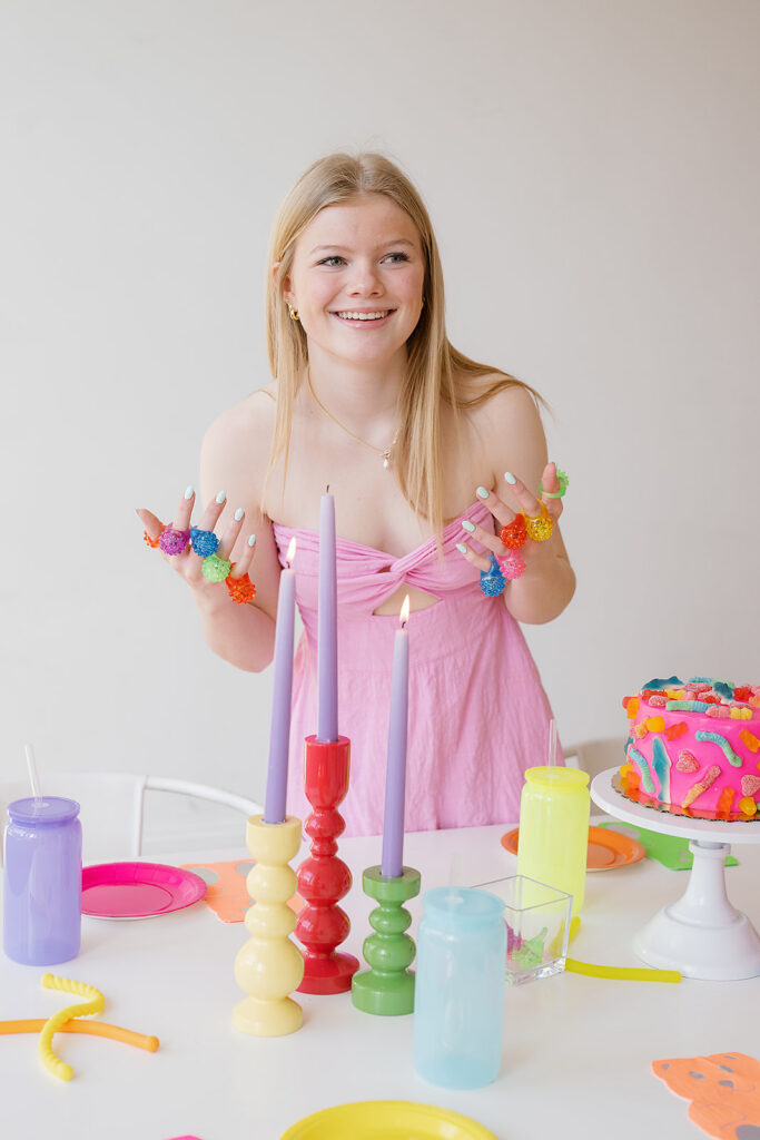A blond girl in a pink strapless dress with gummy like rings on each finger. She is leaning over a table with colorful cups, plates and candles and a gummy pink cake. 