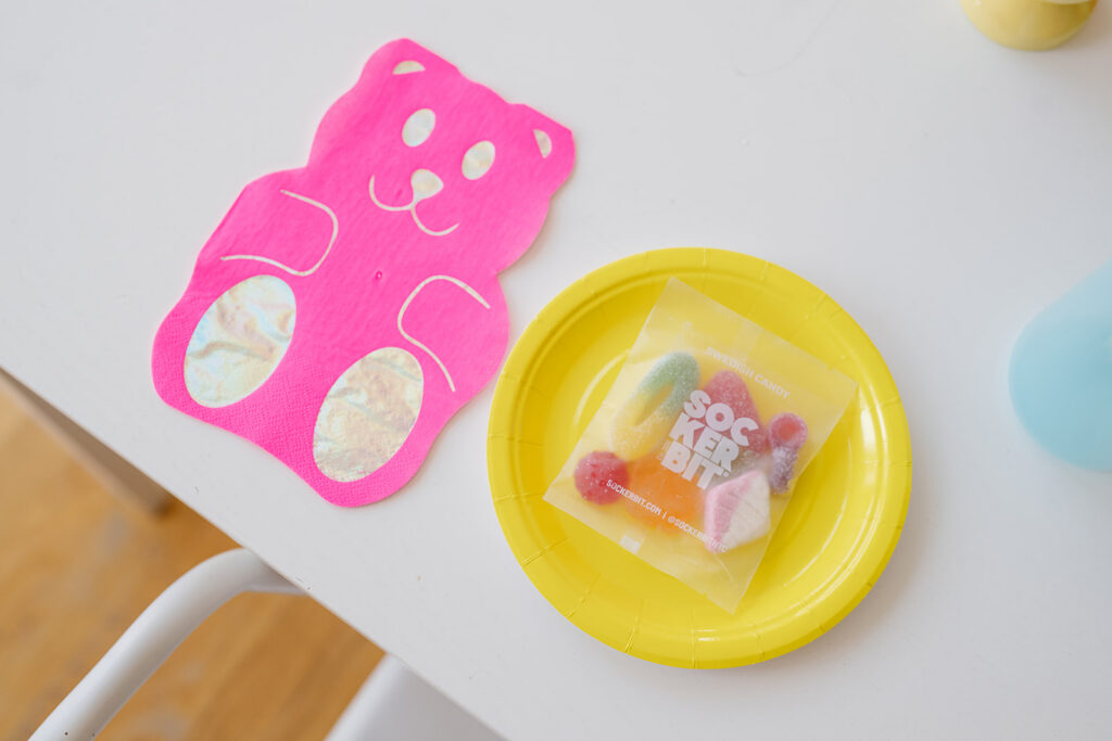 A close up photo of the place setting of a pink gummy bear napkin and yellow plate with a small package of sockerbit gummies on the plate