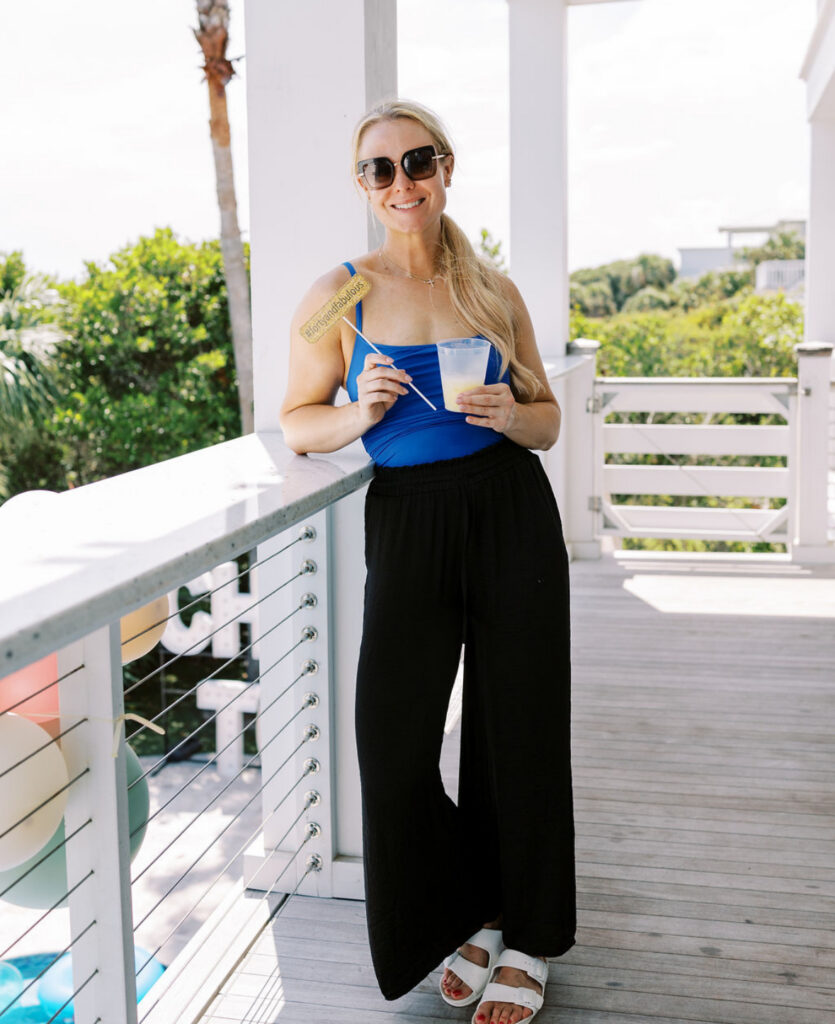A blind haired lady in a blue swimsuit and black pants holding a drink a nd leaning on a balcony