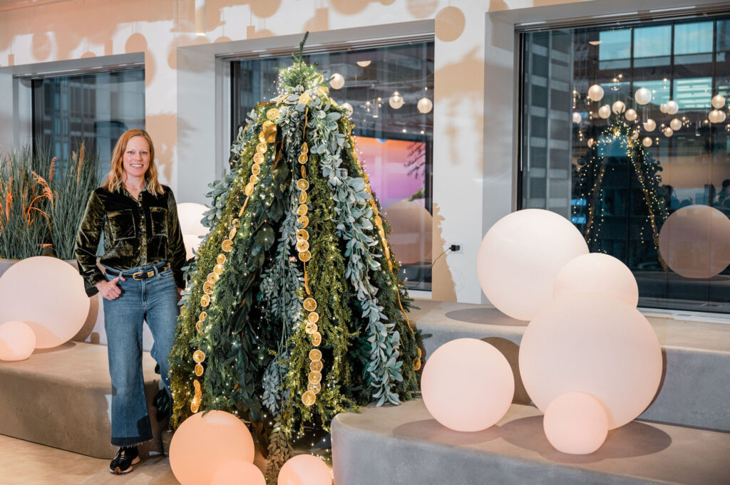 Jessie the founder of WE EVENTS standing next to a heavily decorated evergreen tree. The tree is adorned with lots of different greenery and dried oranges. Large soft white orbs are on the couches next to her. 