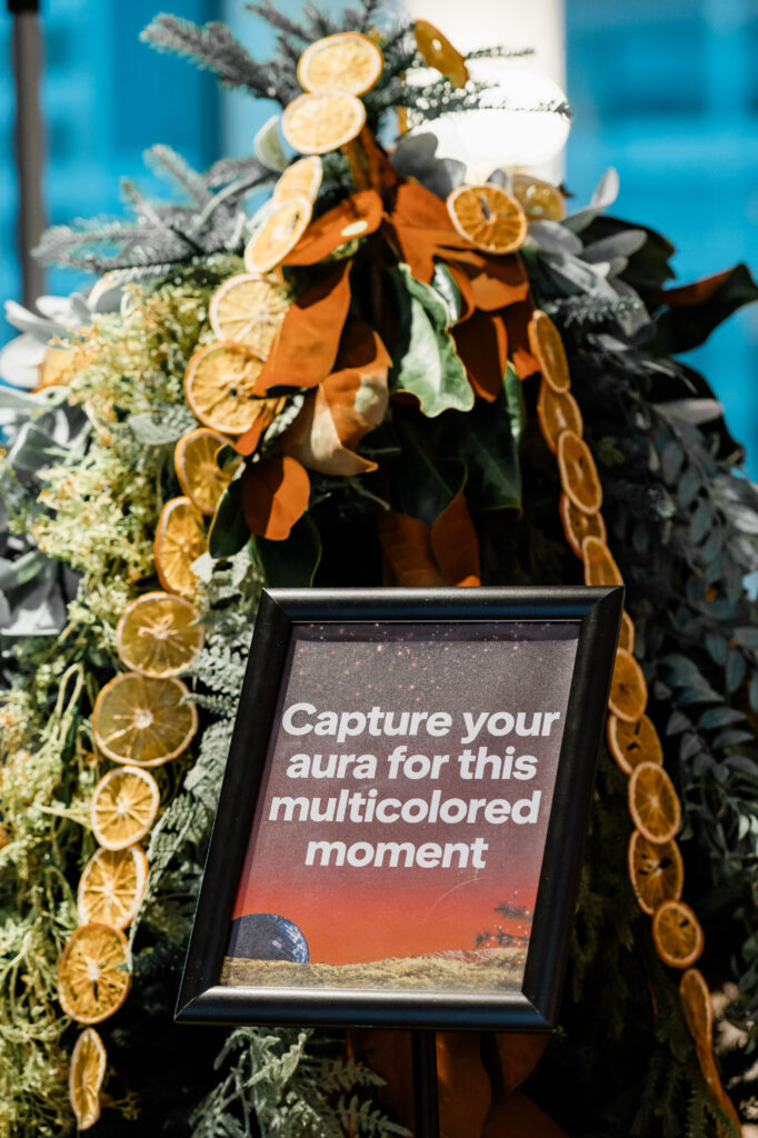 A decorate pine tree with lots of greenery and dried oranges with a sign in front of it saying: capture your aura for the multi colored moment